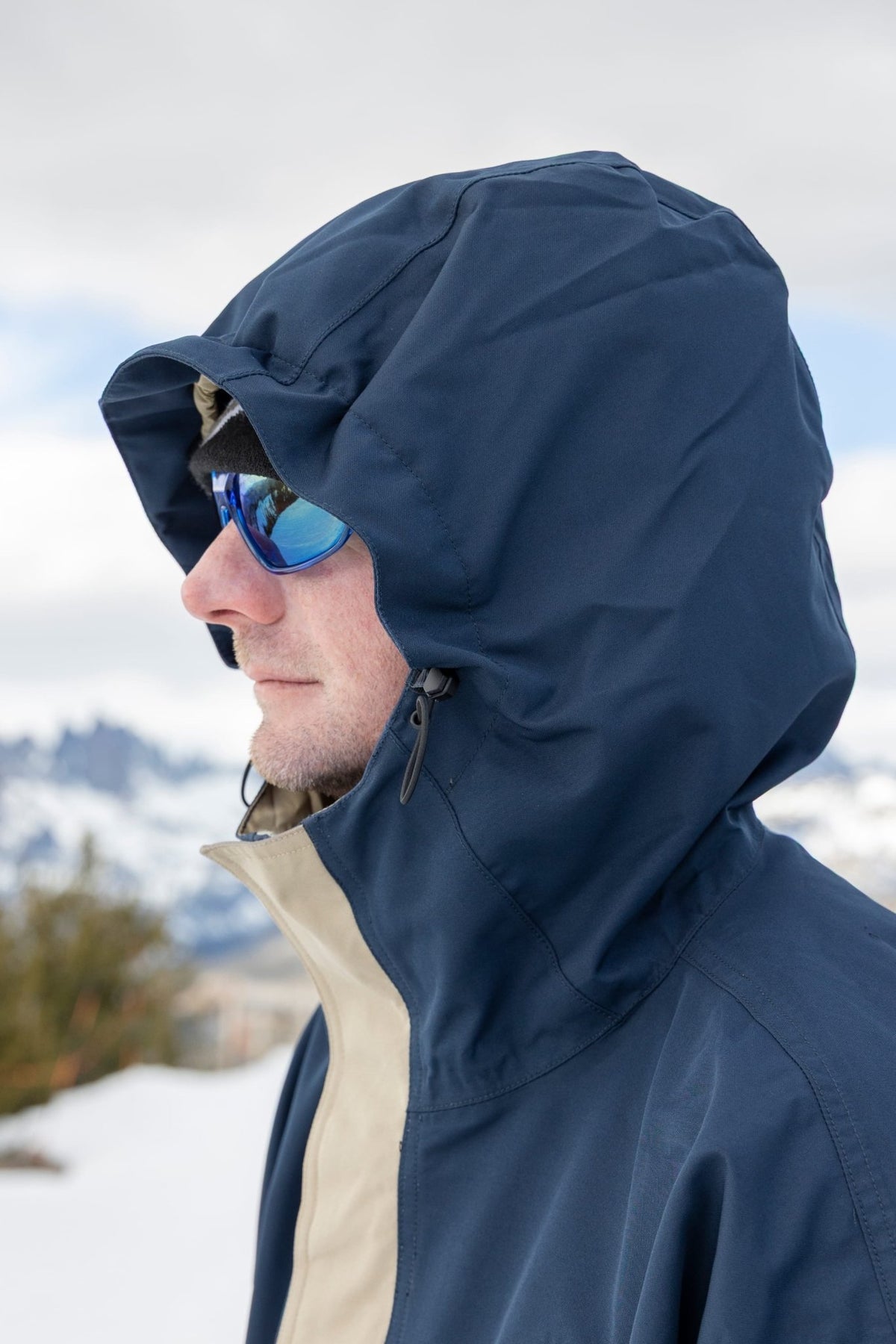 Men's hooded ski jacket with adjustable hood and blue mirrored sunglasses in snowy mountain setting.