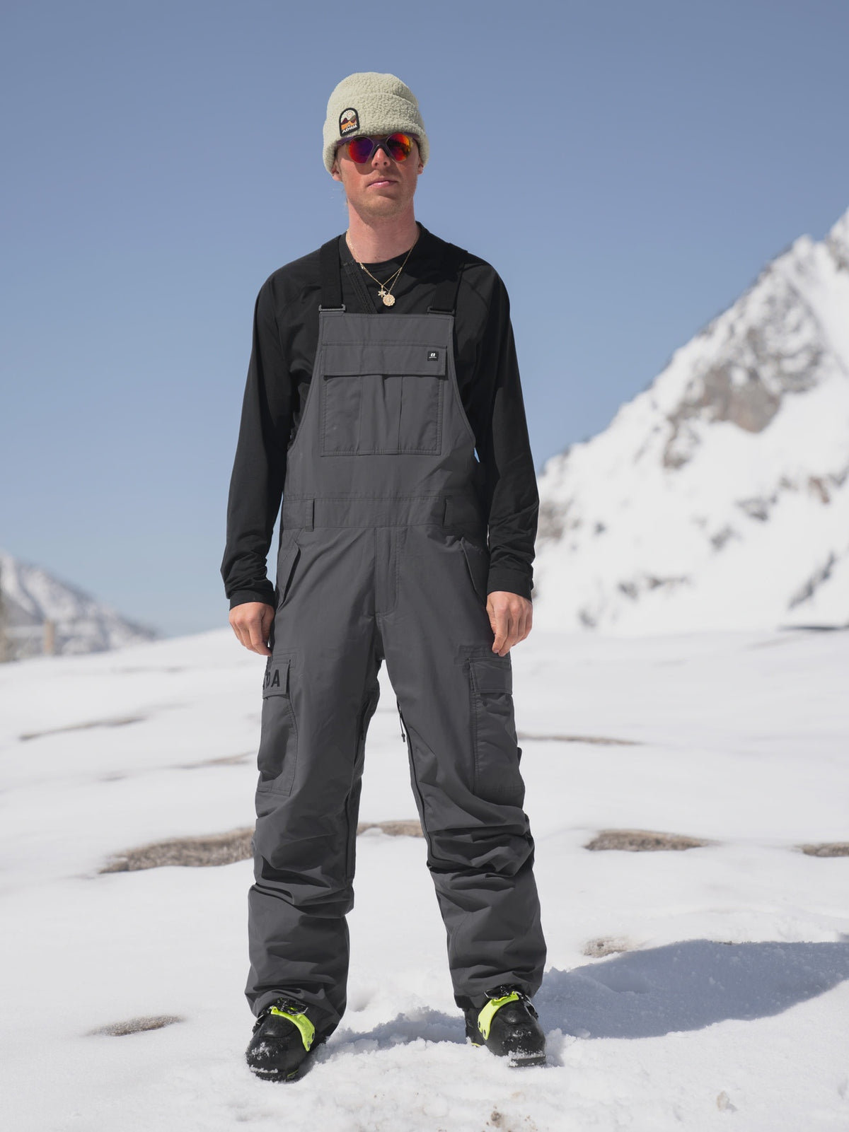 Men's dark gray ski bib overalls with cargo pockets worn over black base layer on snow