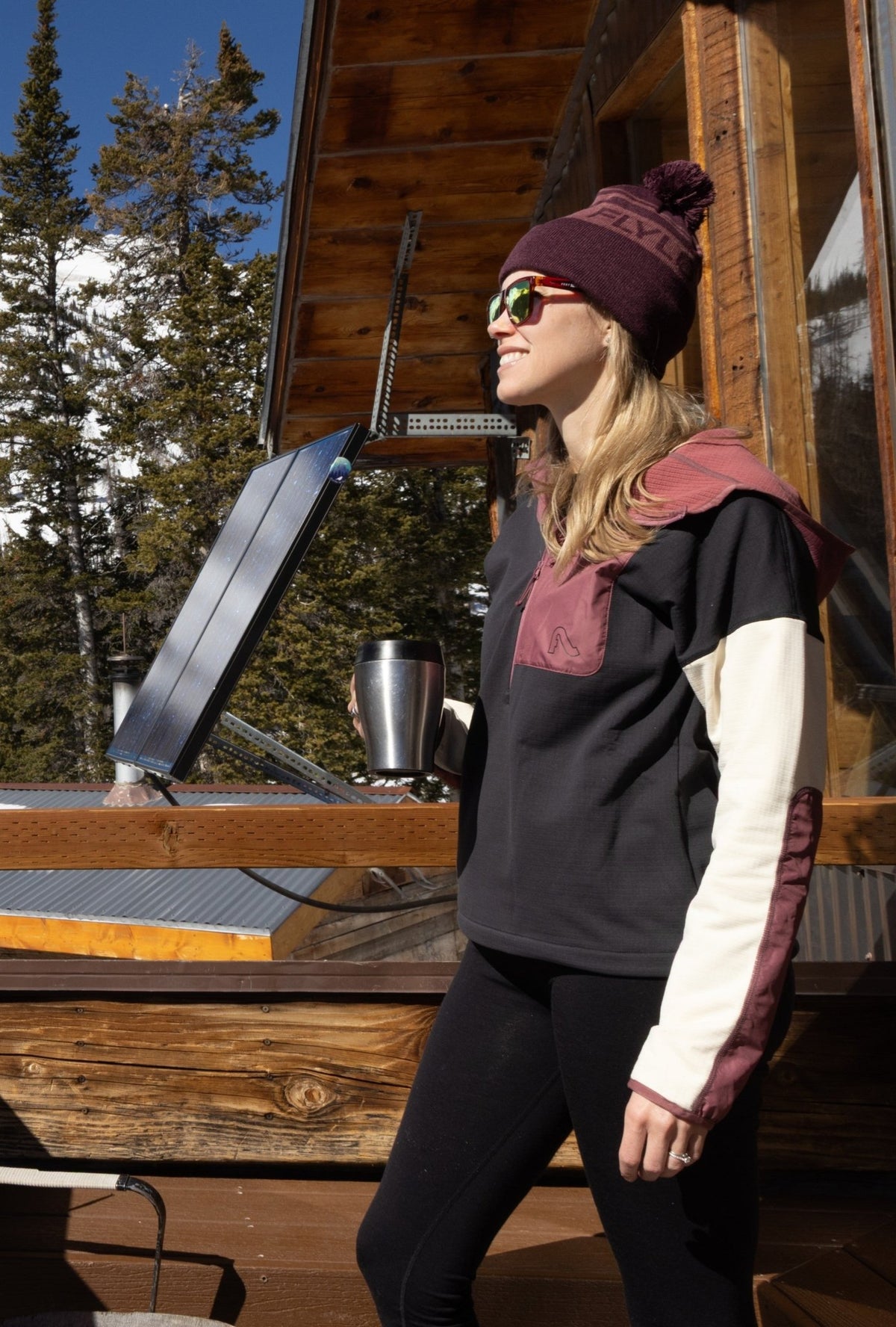Woman wearing Flylow pullover hoodie with colorblock design, standing outside a mountain cabin in winter