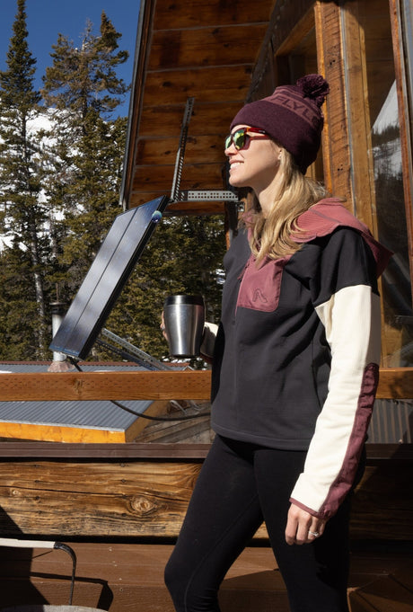 Woman wearing Flylow pullover hoodie with colorblock design, standing outside a mountain cabin in winter