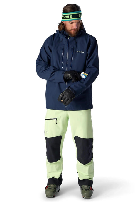 Flylow men's ski jacket in navy blue shown with light green ski pants on male model