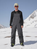 Men's dark gray ski bib overalls with cargo pockets worn over black base layer on snow